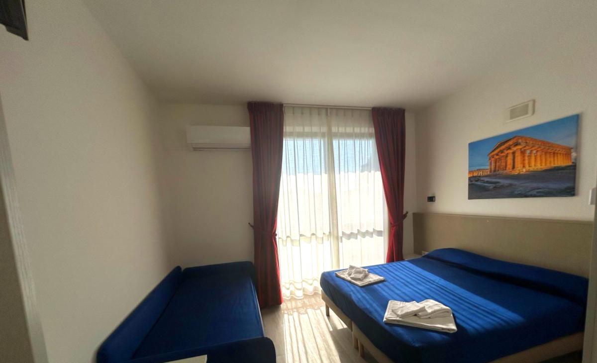 Camera luminosa con letto blu, tende rosse e quadro di tempio antico.