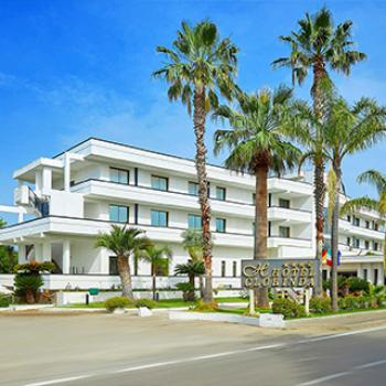 Hotel moderno con palme, facciata bianca e ampi balconi su strada.