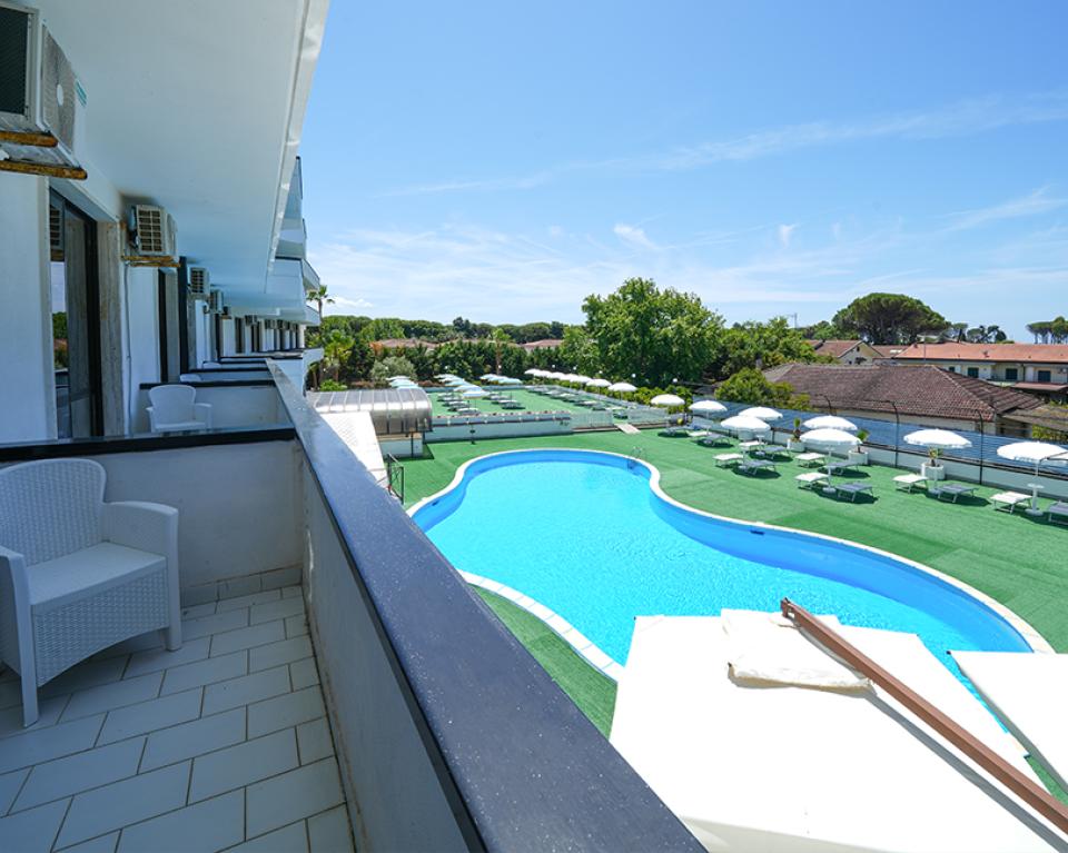 Vista su piscina e giardino da un balcone con sedie bianche.